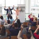 Rear view of diverse business people applauding and celebrating while they are sitting in front of multi-ethnic business executives holding arms up in the air at business seminar in office building