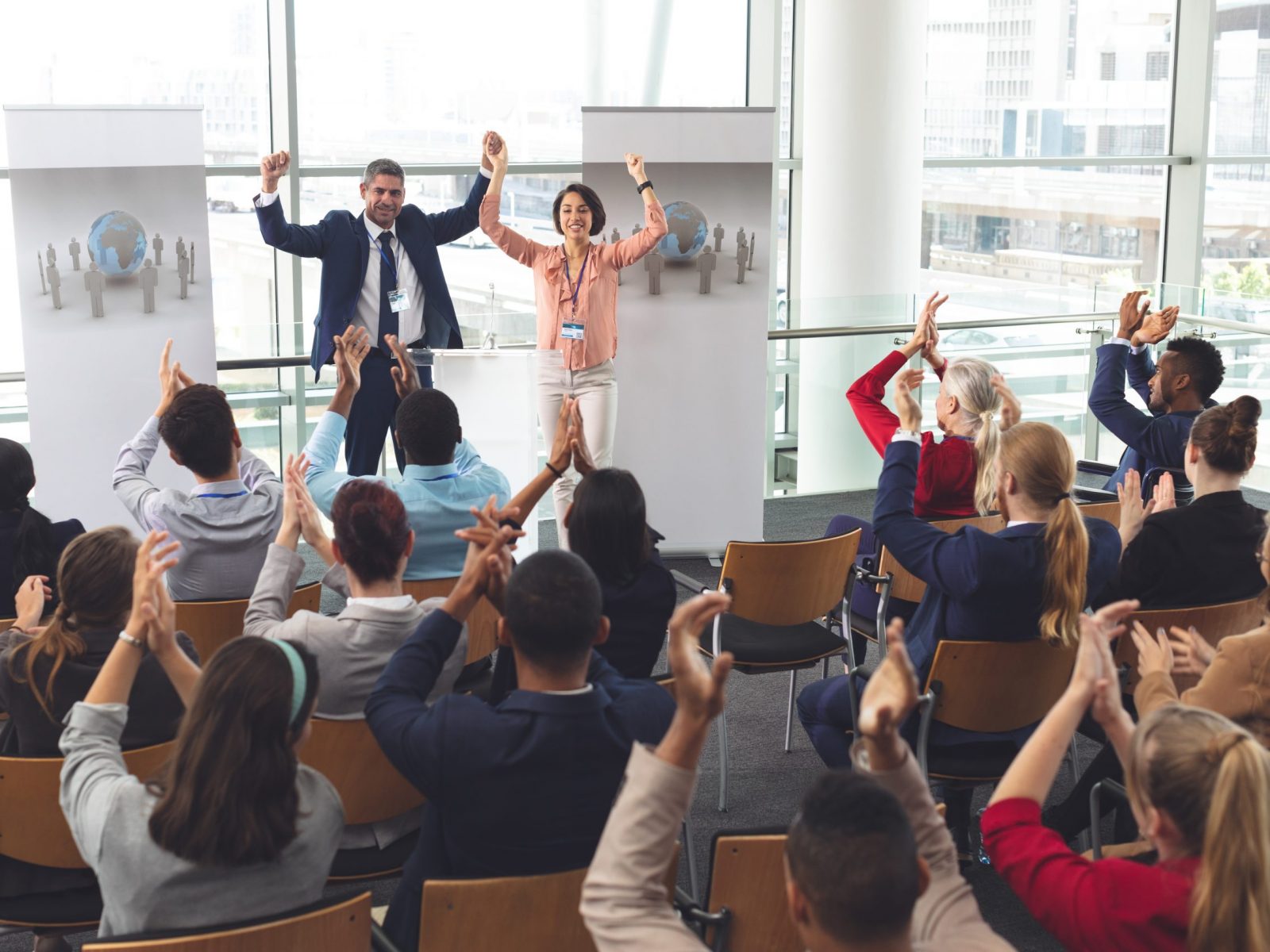 Rear view of diverse business people applauding and celebrating while they are sitting in front of multi-ethnic business executives holding arms up in the air at business seminar in office building